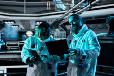 Takım çalışması yapan astronotlar dizüstü bilgisayar kullanarak kıyafet bütünlüğünü doğrulamak için beyin fırtınası yapıyorlar. Yörünge istasyonu meslektaşları konuşuyor, not defteri kullanarak rutin ekipman kontrolü sırasında teçhizat deniyor.