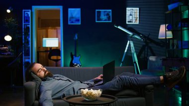 Bütün gün dizüstü bilgisayarda çalıştıktan sonra kanepede uyuyan bitkin bir adam TV izlemeden ve patlamış mısır yemeden önce. Freelancer not defterindeki görevleri çözerken ve film izlerken uykuya dalıyor, kamera B