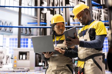 Bilgisayarı ve tableti olan Afro-Amerikan mühendislik ekibi otomatik fabrikada bakım verileri topluyor. Profesyonel teknisyenler akıllı üretim enerji santralinde sektör 4.0 yazılım kullanıyor..