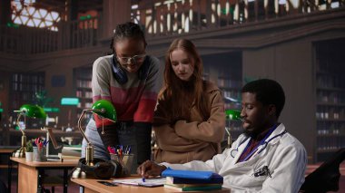 Bir kütüphanedeki çok ırklı öğrenci grubu, tıp kitapları ve materyalleri kullanarak farmakoloji ve mikrobiyoloji alanında uzmanlık kazanmak amacıyla doktorla birlikte bir derse katılıyor. Bilimsel çalışmalar. Kamera B.