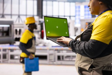 Kadın mühendis akıllı fabrikada yeşil ekranlı dizüstü bilgisayar kullanırken endüstriyel otomasyon dijital izleme ile buluşuyor. Robotik silahlar ve taşıma bantları üretim tesisinde üretim sürecini düzene sokuyor.