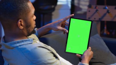 Oturma odasında izole edilmiş bir ekranı olan rahat bir erkek kullanıcı tutma cihazı. Kanepede oturuyor ve telgraf çekiyor. Afrikalı Amerikalı mutlu adam interneti geziyor, dış kaynak kullanıyor. Kamera B.