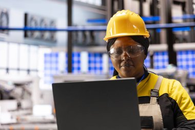 Bilgisayarındaki makine yazılım güncellemelerini inceleyen kadın uyum mühendisi. Afrikalı Amerikalı endüstriyel teknisyen akıllı fabrikada yenilikçi altyapılar geliştirmek için çalışıyor..