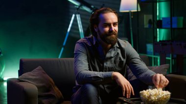 Evde boş zamanlarında televizyonda kablolu yayın izlerken patlamış mısır yiyip gülümseyen bir adam. Oturma odasındaki kanepede oturup abur cubur yiyor ve televizyon dizisi, kamera A 'ya bakıyor.