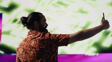 Genç kaygısız adam, modern DJ kültürünün, medya etkisinin ve dijital sanatın performansını temsil eden eğlenceli neon ışıklar ve ritimlerle çevrili selfie ve videolar çeker. Kamera A.