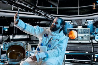 Fütürist uzay aracının içinde uzay elbisesi giyen astronot akıllı telefonuyla selfie çekiyor. Uzay yolculuğu sırasında teçhizat takan gülümseyen uzay adamı cep telefonuyla fotoğraf çekiyor