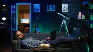 Televizyon izleyip patlamış mısır yemeden önce bütün gün dizüstü bilgisayarda çalıştıktan sonra kanepede uyumamak için çırpınan uykucu adam. Defterdeki görevleri çözerken ve film izlerken uyuyakalan kişi