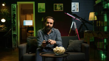 Apartmandaki kanepede oturan oyuncu oyun platformunu kullanarak online çok oyunculu bir turnuvaya katılıyor. Ev sinemasında oyun oynayan adam kontrolörle oyun oynuyor, patlamış mısır yiyor, kamera B