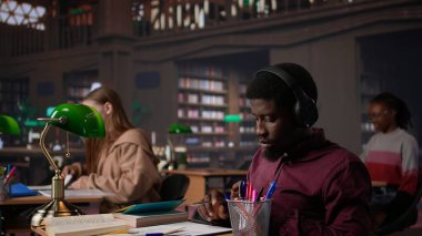 Ortam müziği dinleyen, kulaklıkla müzik dinleyen ve okul projelerine odaklanan bir kitaptan veri toplayan erkek öğrenci. Okul ödevini bitirmek için ortam müziğinin tadını çıkarıyorum. Kamera B.