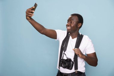 Siyah erkek fotoğrafçı mavi arka plandaki bir stüdyoda cep telefonunu güvenle tutarken bir selfie çekiyor. Pantolon askısı takıyor, boynunda bir kamera var, şık ve modern bir görünüm yayıyor..