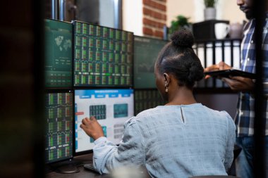 Kadın borsa danışmanı, meslektaşıyla yaptığı yatırımlardaki canlı güncellemeleri açıklarken bilgisayar ekranını işaret ediyor. Siyahi bir kadın, çok monitörlü masada oturmuş, ön x piyasasını analiz ediyordu..