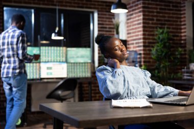 İş yorgunluğu çeken Afrikalı Amerikalı bir kadın dizüstü bilgisayarındaki verileri incelemeye ara vererek boynuna masaj yapıyor. Gece vardiyası, gece vardiyalarının yoğunluğunu vurguluyor..