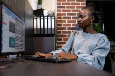 Geç saatlere kadar masasında kalan siyahi iş kadını pek çok ekrandaki satış grafiklerini izliyor. Onun tuğla duvar ofis ayarı çalışma ortamına modern bir dokunuş katıyor..