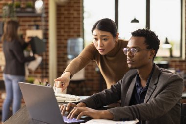 Kadın analist, Afro-Amerikan bir iş arkadaşına pazar analizini açıklamak için laptop ekranını işaret ediyor. Siyahi adam, Asyalı menajerin sunumdaki mali hataları ele alışını dikkatle dinliyor.