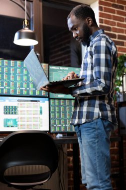 Afrikalı Amerikalı yatırımcı, loş ışık odasındaki borsa grafikleri ve ekonomik eğilimleri, dizüstü bilgisayar ve yatırım büyümesini izlemek için çoklu ekran kurulumu kullanarak inceliyor. Modern teknoloji ofiste kullanılıyor..