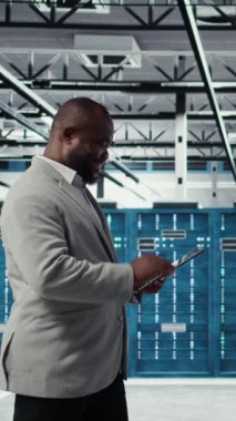 Veri merkezinde sistem istikrarı için yazılım güncellemelerini yönetmek için tablet kullanan dikey video programcısı. Sunucu odasında Afrikalı Amerikalı bir adam performansı cihazla optimize ediyor, yapılandırıyor.