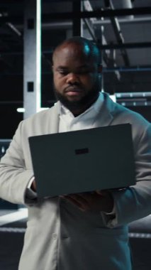 Dikey video veri merkezi IT uzmanı ekipman donanımlarının performans gösterge panellerini inceliyor. Sunucu odası yöneticisi sistemleri denetliyor, laptop ekranındaki altyapı algoritmalarını inceliyor, kamera A