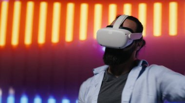 Neon ışıklı bir gece sahnesinde modern bir adam VR yüksek teknoloji gözlüğü kullanıyor, siber punk stilini somutlaştırıyor ve sanatsal vizyon yoluyla dijital ilham veriyor. Fütürist estetiğin parlak yansımaları. Kamera A.