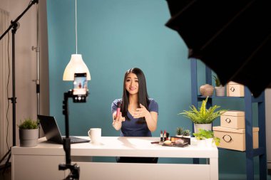Güzellik içeriği yaratıcısı stüdyoda cep telefonuyla video kaydediyor, seyircisini kozmetik bir incelemeyle meşgul ediyor. Sahne mavi arkaplan, aydınlatma ekipmanı ve tripodun üzerindeki mobil aygıtı içeriyor.