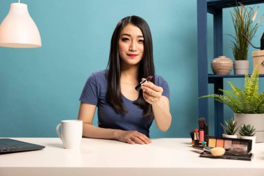 Masada oturan Asyalı güzellik uzmanı makyaj dersini çekiyor. Kadın vlogger bir ruj tutuyor, kameraya sunuyor ve abonelerini uzman kozmetik tavsiyeleriyle meşgul etmek için bir video kaydediyor..