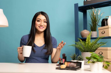 Genç Asyalı vlogger canlı bir seans sırasında izleyicileriyle konuşurken bir fincan kahvenin keyfini çıkarıyor, mavi arka planı olan bir stüdyoda çekim yapıyor. Güzellik etkileyicisi aboneleri için içeriği kaydediyor..