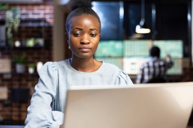 Kadın finans analisti geceleri masasında oturup kişisel bilgisayarındaki borsa bilgilerini kontrol ediyor. Arka planda parlak monitörleri olan teknoloji meraklısı bir çalışma istasyonu görülüyor.