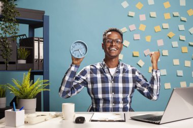 Elinde saatle mutlu bir işadamı, proje sunumunu son teslim tarihinden önce bitirdiği için kutlama yapıyor. Neşeli erkek menajer heyecanlı bir şekilde yumruğunu kaldırıyor, günün sonunda kendini harika hissediyor..