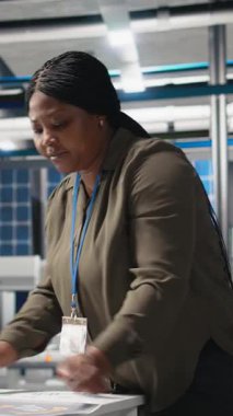 Dikey Afrikalı Amerikalı kadın uzman güneş panelleri üretim hattını inceliyor, bir fotovoltaik fabrikasında kalite kontrolü yapıyor. Güneş enerjisi yaratmak için veri raporları ve içgörü analiz ediliyor. Kamera.