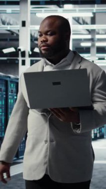 Yapay zeka otomasyon kodlarını incelemekle görevli dikey video veri merkezi sistem yöneticisiName Afrika kökenli Amerikalı bir adam sunucu merkezine girip ekipman teçhizatı ve kamera A 'yı kontrol ediyor.