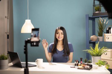 Mobil bir cihazda güzellik etkeni dalgaları, stüdyo masasında kozmetik incelemeye hazırlanıyor. Asyalı kadın vlogger çevrimiçi izleyicileri için üç ayaklı akıllı telefon kullanarak bir makyaj dersi kaydediyor.