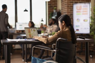 Tekerlekli sandalyedeki profesyonel kadın, tuğla duvardaki ofiste dizüstü bilgisayarını kullanıyor, araba kazasından sonra işleri hallediyor. Asyalı bayan girişimci modern çalışma alanında iş projesi sunumunu gözden geçiriyor.