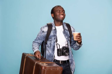 Bir elinde bavul, boynunda da kamerayla gezintiye çıkmaya hazırlanırken uyanık kalmak için kahve içtiği için heyecanlı bir seyahat videosu kaydedicisi. Bavullu ve sırt çantalı siyah erkek fotoğrafçı, gülümsüyor..