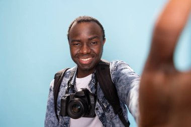 Gülümseyen fotoğrafçı stüdyodaki video kayıt kanalı için içerik kaydediyor. Boynunda DSLR kamerasıyla, izole edilmiş arka planda neşeyle selfie çeken Afrikalı Amerikalı bir adam..