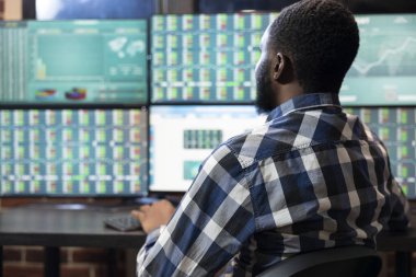 Masadaki genç gece tüccarı ofisteki birden fazla monitörde ön market grafiklerini analiz ediyor. Afrika kökenli Amerikalı profesyonel bilgisayar ekranlarının önünde oturup gerçek zamanlı hisse senedi hareketlerini izliyor..