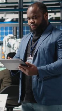 Bir güneş enerjisi üretim tesisinde çalışan dikey video Afrikalı Amerikalı teknisyen şirket denetimi ve ekipman üretimini yansıtmak için takım elbise giymiş operasyonları değerlendiriyor. Temiz enerji. Kamera.