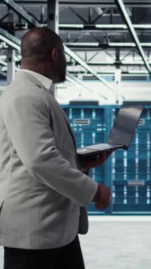 Sistem istikrarını yükseltmek için dizüstü bilgisayar kullanarak gülen dikey video veri merkezi programcısı. Neşeli Afro-Amerikan sunucu merkezi bilişim uzmanı kodlama, cihaz ile performansı optimize etme, ayarlama ekipmanları, kamera