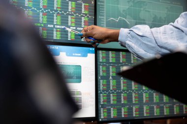 Afrikalı Amerikalı girişimci kadın, dijital çalışma alanındaki anahtar pazar hareketlerini vurgulamak için bir kalem kullanarak birden fazla monitörden bilgi alışverişinde bulunuyor. Kadın eli bilgisayar ekranını işaret ediyor.