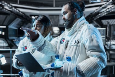 Bir grup astronot dizüstü bilgisayar kullanarak kıyafet bütünlüğünü doğrulamak için birlikte çalışıyor. Yörünge istasyonu çalışanları sohbet ediyor, rutin ekipman kontrolü sırasında not defteri kullanarak teçhizat deniyor.