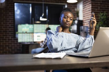 Bir elinde kalemle yorgun düşmüş kadın girişimci, uzun saatler süren borsa analizinden bunalmış bir şekilde loş bir ofiste boynunu ovuyor. Bilgisayarı açık kalmış, ticaret çizelgelerini gösteriyor..