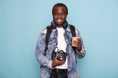 Kameralı ve sırt çantalı Afro-Amerikan bir adam sıcak bir içeceğin tadını çıkarıyor ve izole arka planda duruyor. Kot ceketli neşeli fotoğrafçı elinde kahve fincanı, çekim gününe hazır..