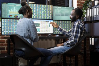 Masada yan yana çalışan iki Forx tüccarı, geceleri profesyonel ofis ortamında çok ekran iş istasyonundaki borsa eğilimlerini izliyorlar. Siyahi kadın ve erkek yatırım stratejilerini tartışıyor.