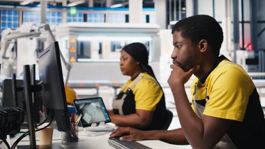 Siyah mühendis otomatikleştirilmiş fabrikada güneş yazılımı ayarları üzerinde çalışarak üretim sürecini optimize ediyor. Dijital araçlar ve endüstriyel robotlar kullanılarak optimal sistem yapılandırmasını sağlar. Kamera B.