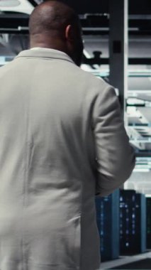 Dikey video veri merkezi sistem yöneticisi tablet ekran kullanarak altyapı teşhislerini kontrol ediyor. Yüksek teknoloji sunucu odasında yürüyen bilişim uzmanı, sistem denetimi sırasında veri tablolarını inceliyor