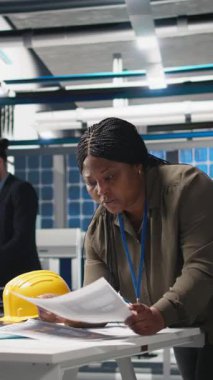 Güneş enerjisi alanındaki dikey video Afrikalı Amerikalı endüstriyel müfettişi kağıtlardaki verileri inceler, üretim hattını ve üretim sürecini inceler. Bitki performansını kadın uzman denetliyor. Kamera A.