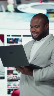 Dikey videodaki gülümseyen kişi dizüstü bilgisayar kullanıyor ve video yayın hizmetlerinin içeriğinden keyif alıyor. Neşeli adam dijital arkaplan önünde VOD medya yayınlarını izliyor