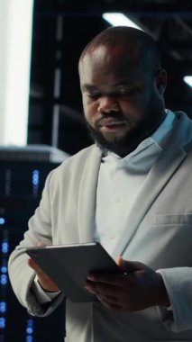 Veritabanı kontrol listesindeki tabletlere bakan dikey video uzmanı. Afrikalı Amerikalı bir adam sunucu çiftliğinde yürüyor, cihaz üzerindeki dökümantasyon dosyalarını okuyor, kamera B