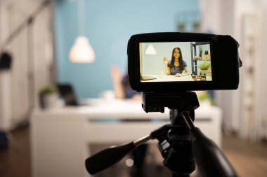 Tripodun üzerindeki dijital kamera mavi arka planı olan bir stüdyoda Asyalı bir güzellik blogcusunu kaydediyor. Kadın içerik yaratıcısı arka planda bulanıklaşıyor ve canlı yayına bağlanıyor..