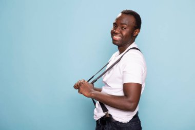 Afrikalı Amerikalı bir adam pantolon askılarını bir stüdyo resmine göre ayarlıyor. Kendine güveni ve parlak gülümsemesi izole edilmiş arka planda güçlü bir görsel etki yaratıyor..