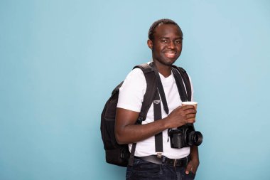 Sırt çantalı, elinde kahve fincanıyla kameraya bakan Afrikalı Amerikalı bir adam. İzole edilmiş arka planda duran erkek fotoğrafçı, teçhizat hazır ve sıcak içecek elinde, fotoğraf çekimine hazır..