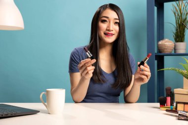 Farklı olduklarını açıklarken iki ruj gölgesini tutan ve karşılaştıran kadın vlogger 'ın portresi. Sosyal medya etkileyicisi mavi arka plan stüdyosunda çekim yapıyor, yeni makyaj ürünlerini gözden geçiriyor..